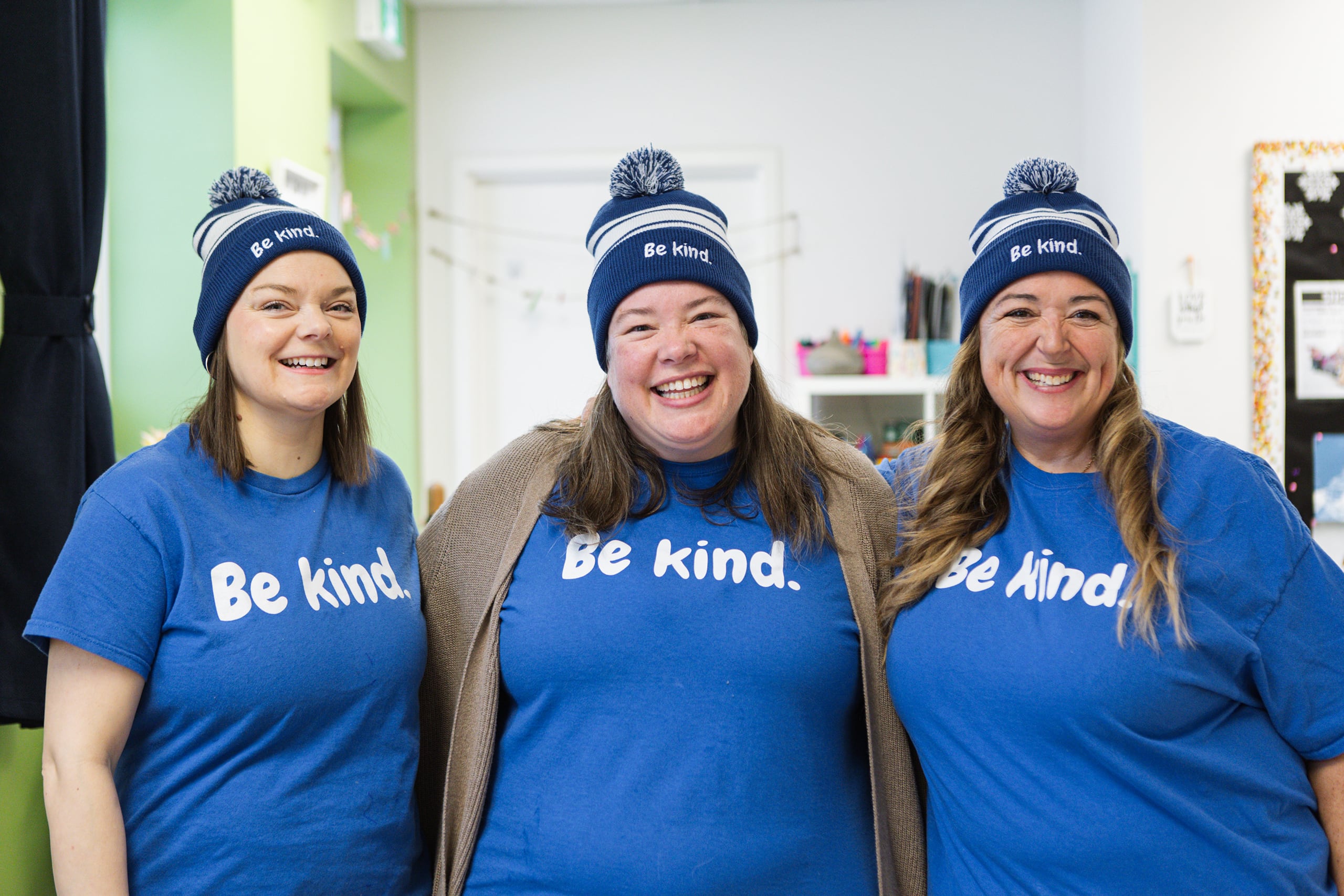Three Kanata Academy staff members smiling and wearing matching blue "Be kind." toques