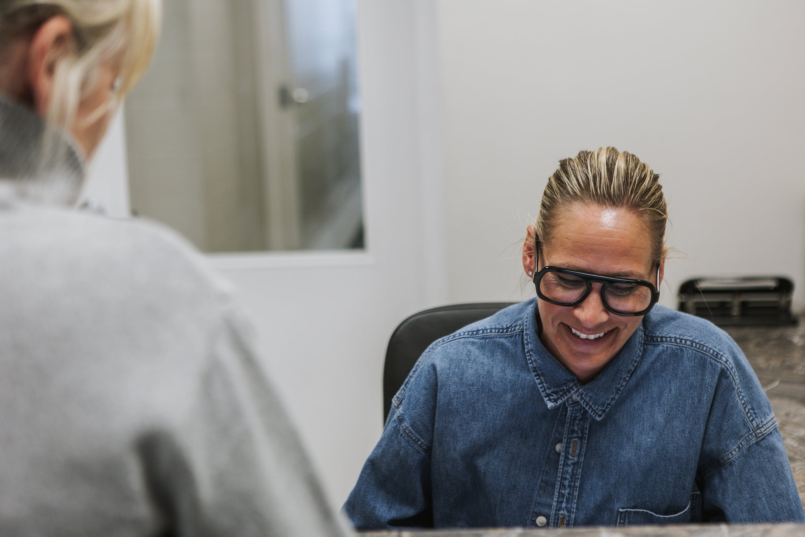 Smiling adult with glasses and blonde hair in a denim jacket, photographed during a meeting