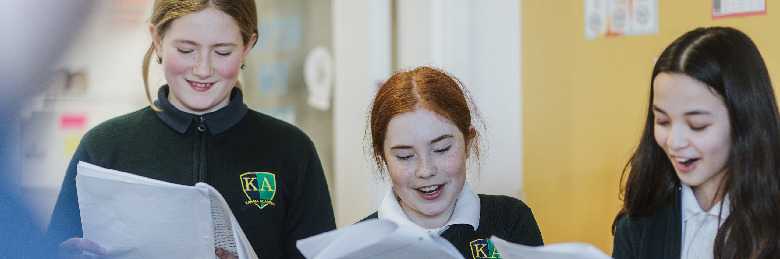 Three junior students in Kanata Academy uniforms reading together and smiling